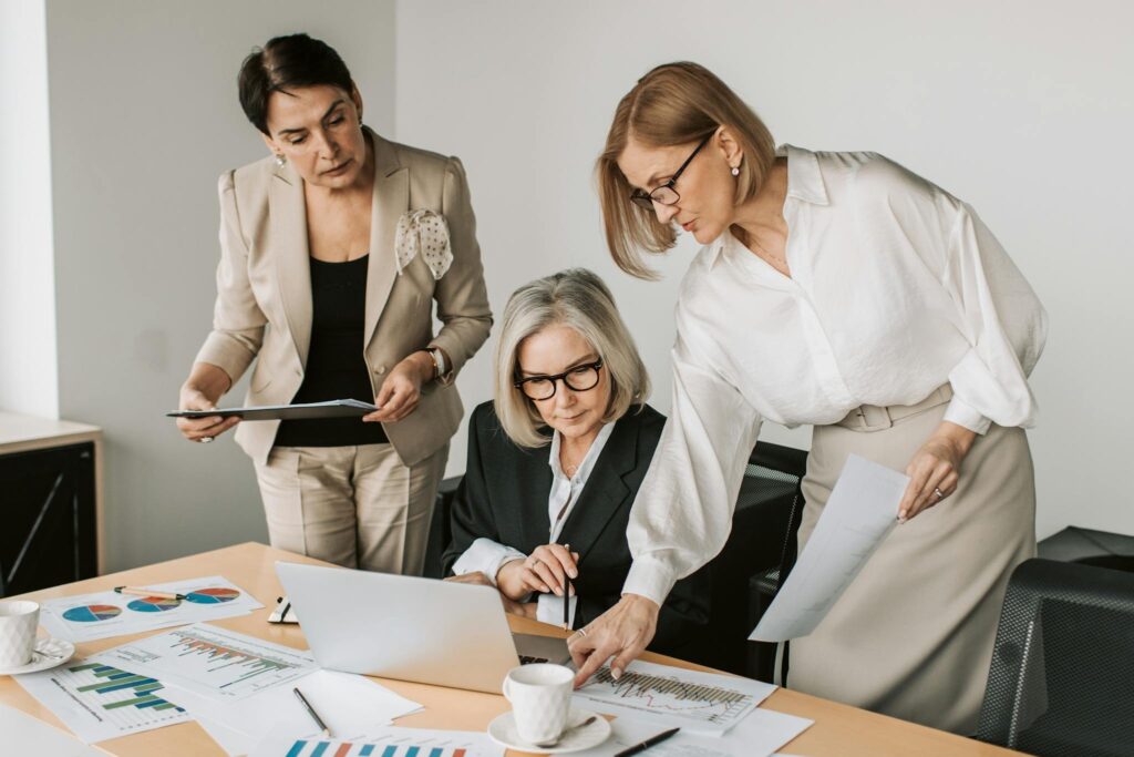 Three senior businesswomen in an office working together on a laptop and documents.